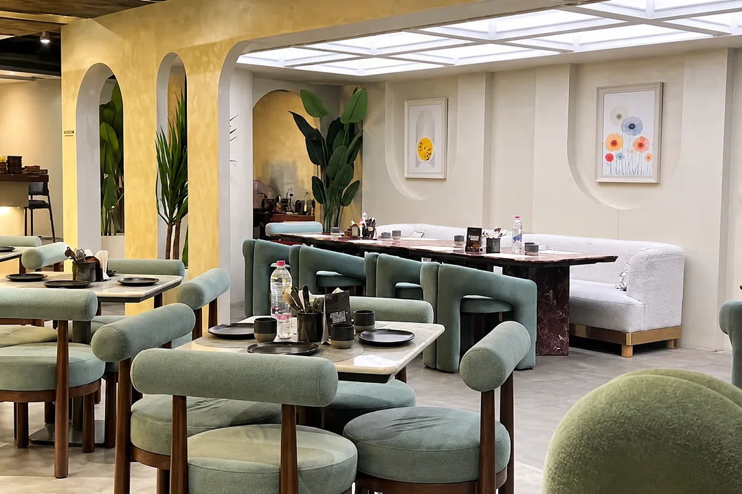 Spacious main dining hall with sage green upholstered chairs, marble tables, long ivory curved banquette, skylight ceiling coffer, and warm plaster arches.