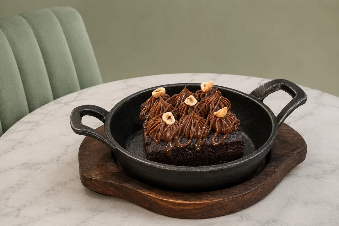 Warm decadent chocolate fudge brownie with piped hazelnut ganache served in a cast iron skillet on a dark timber trivet.
